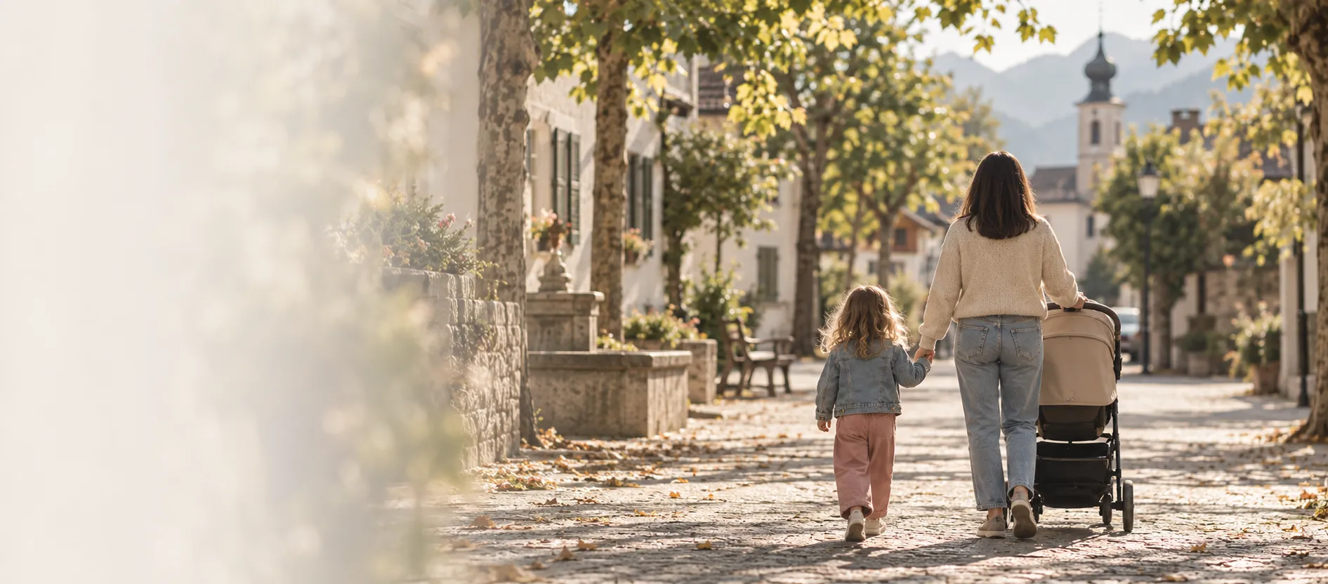 A mother walking with a child and a pushchair along a sunlit street in an Alpine town